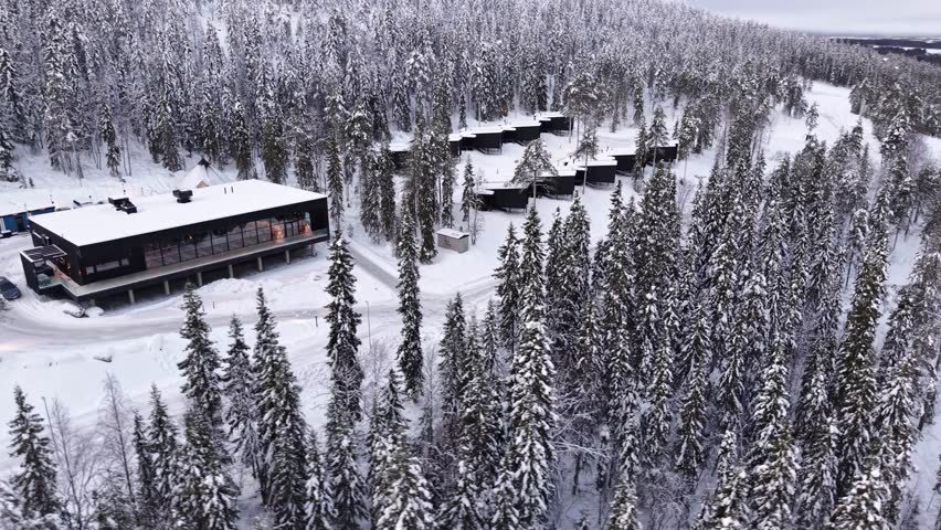 Winter hotel surrounded by pine forest covered in snow, Glass cabins, chalets near Santa Claus village, Lapland, drone shot