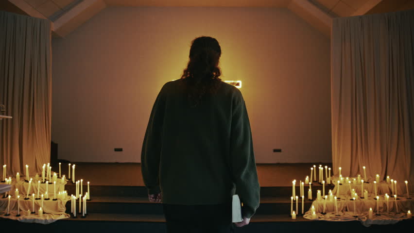 Young man carrying a bible walking towards a glowing cross in a dark church with burning candles