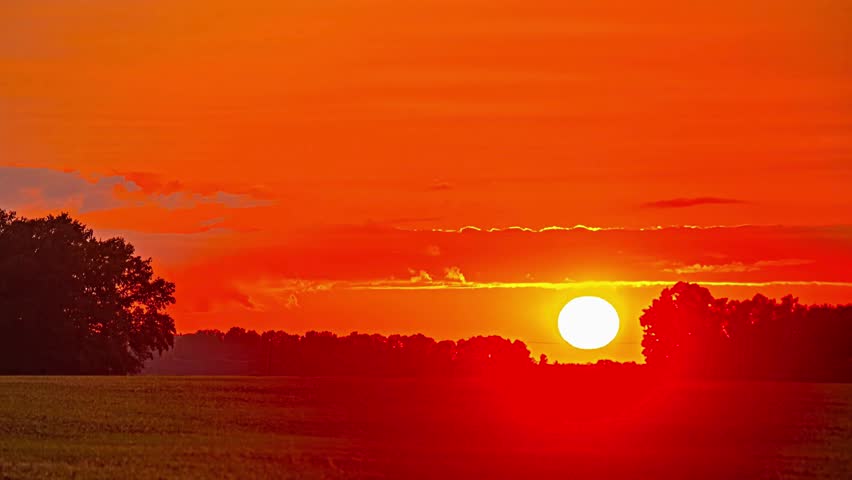 A warm orange glow fills the sky as the sun sets over serene rural fields. Captured in panoramic time-lapse, this landscape highlights peaceful farmland, rolling hills, and natural beauty.
