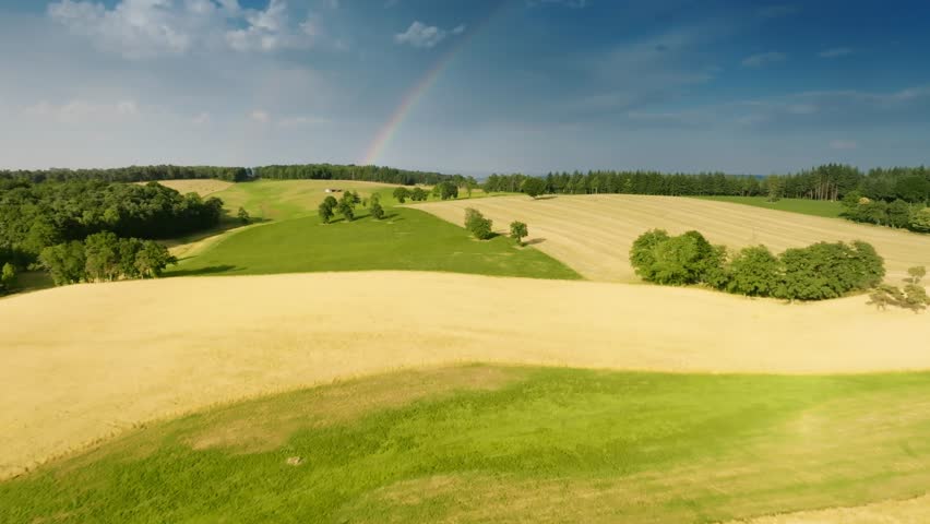 Low aerial drone flight gliding over a golden wheat field towards green trees. A subtle rainbow arcs in the blue sky above the French countryside landscape.