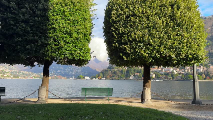 Beautiful view of Como lake, Italy. Promenade with green trees in Como town. Bench on the shore of lake between the trees. Beautiful landscape at sunny day
