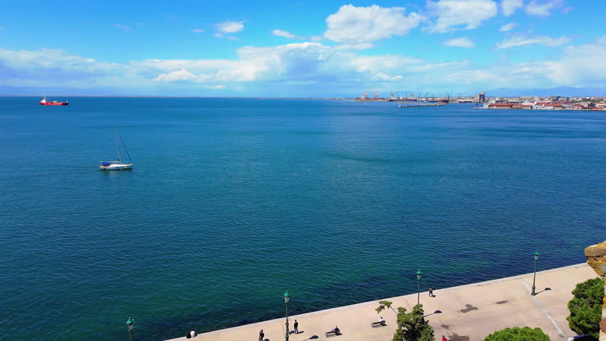 view of a Greek seaside promenade with people walking along the waterfront and boats on the calm blue sea under a bright sky.