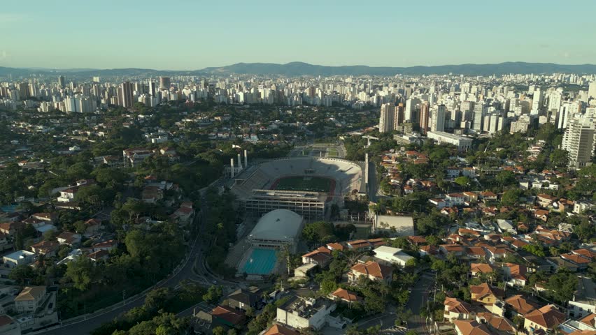 Aerial traveling shot over Pacaembu Stadium. The iconic sports arena, an outdoor swimming pool, and the dense urban horizon of São Paulo.