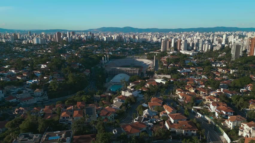 Aerial tilt-up of Pacaembu Stadium. The iconic soccer field, athletic complex, and the dense residential and high-rise skyline of São Paulo at dawn.