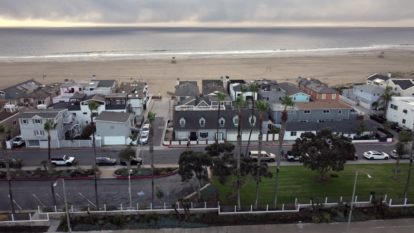 High-angle aerial shot of luxury beachfront homes in Newport Beach, California under cloudy skies. Rainy weather softens the coastal landscape, reflects a slower pace along the Orange County shoreline