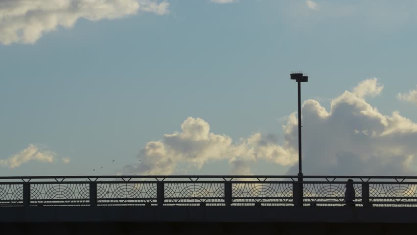 Silhouetted figures of people walking across a bridge against a bright blue sky with fluffy white clouds during late afternoon