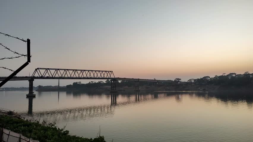 Scenic view of a railway bridge over a calm river at sunrise, reflecting on the water with vibrant sky and lush riverbank vegetation.