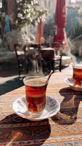 Tea steaming process in Istanbul early morning, two transparent glasses on outdoor cafe table, sunlight highlighting vapor, cold atmosphere visible, blurred urban background. Taken from hand.4k