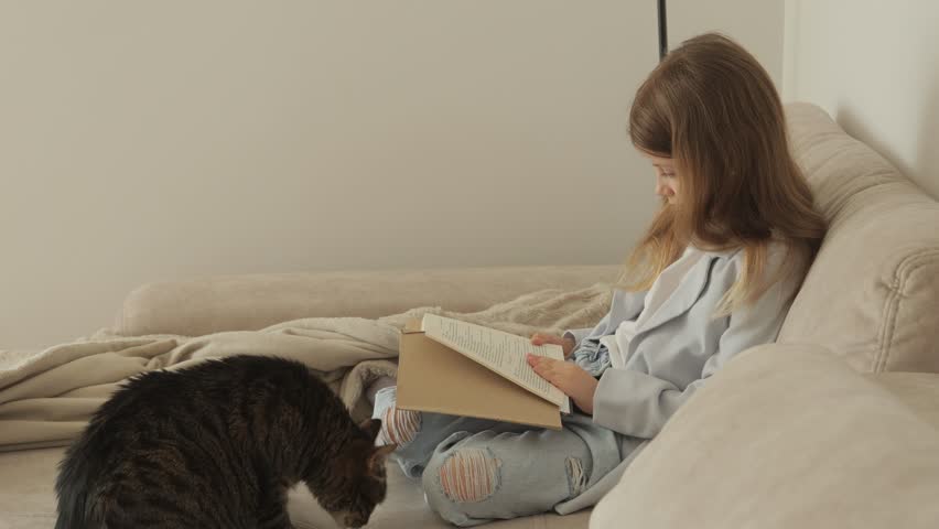 A little girl sits on a beige sofa reading a book with her pet tabby cat sitting beside her. The tabby cat watches her as she reads.