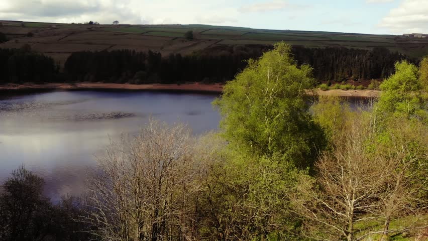 Trees and farmland surround lake in Peak District National Park blue sky landscape wide zoom drone aerial selective focus