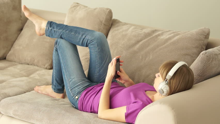 Young woman lying on couch with phone and listening to music
