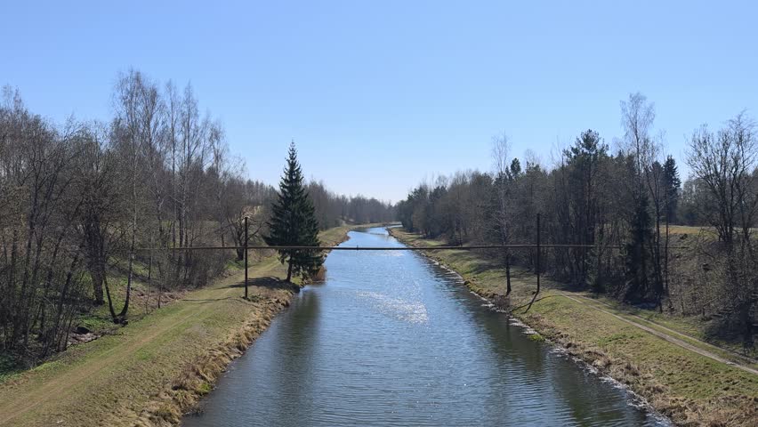 The river flows between grassy banks with dirt roads. Birch, willow, spruce and pine trees grow nearby. A pipeline is installed above the river Ripples on high water. Sunny spring weather and blue sky