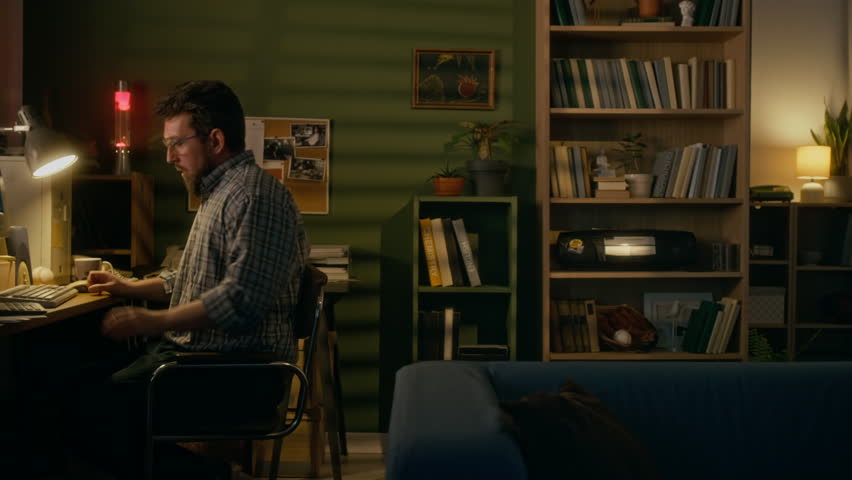 Full side shot of young Caucasian man sitting at desk at home, working on retro PC computer with CRT monitor, typing command and stretching in chair, while connecting to internet using landline modem