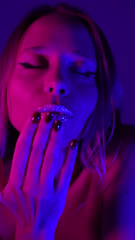 Young blonde woman blowing an air kiss toward the camera, glitter lips glowing in pink and blue neon light. Sensual feminine beauty, soft expression, elegant mood, modern aesthetic, vertical video
