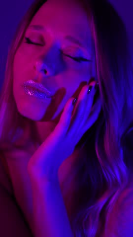Young woman gently touching and stroking her face with closed eyes, glitter lips glowing in pink and blue neon light. Sensual skincare ritual, feminine softness, beauty concept, elegant mood, vertical