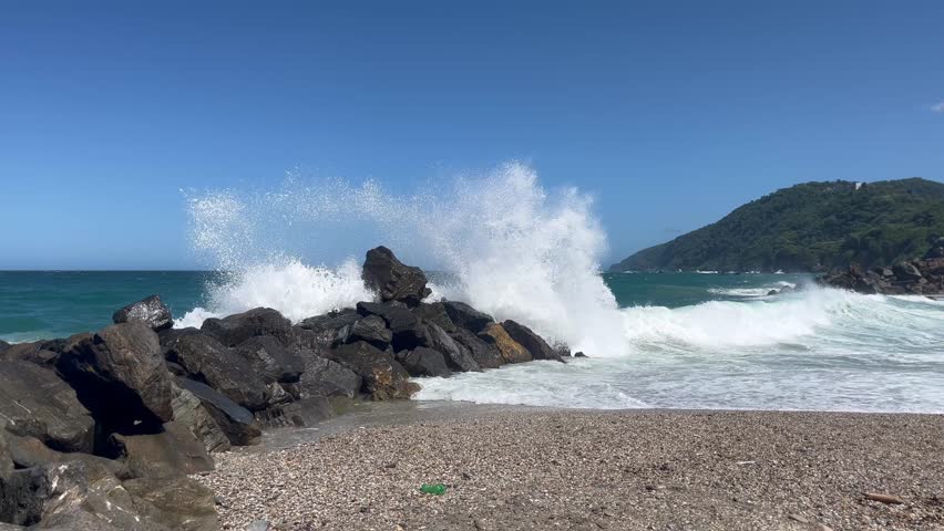 Scenic beach landscape with dark jagged rocks and crashing ocean waves under a clear blue sky