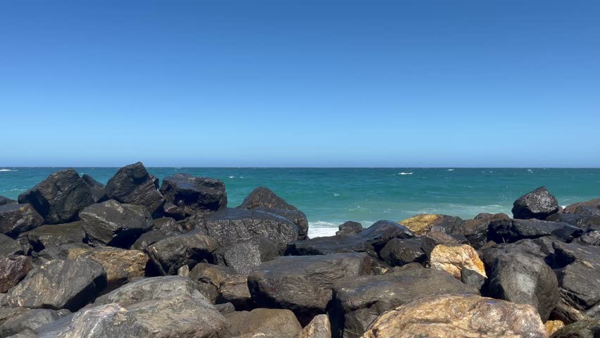 Large dark coastal rocks and boulders on the shore with blue ocean and clear sky background