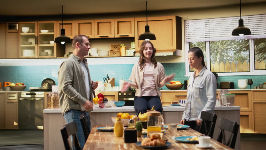 Family in the kitchen dancing and singing during breakfast meal, parents acting goofy and making their kids laugh and have fun. Mom and dad bonding with their children at home. Camera A.