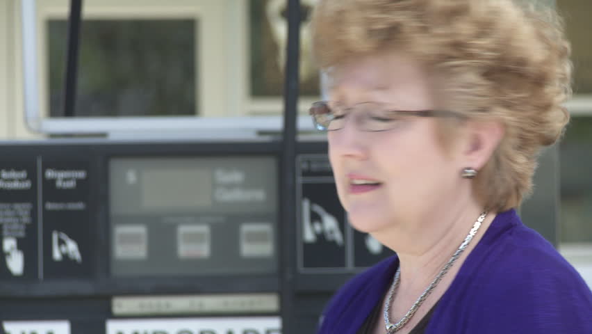 Close up on a worried woman with gas pump and service station in the background.