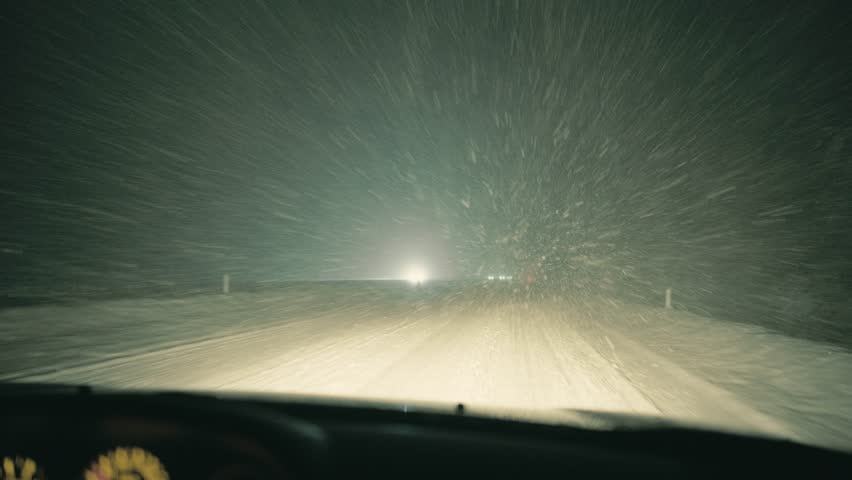 Point of view night driving filmed from inside a car during a snowstorm on a motorway. Poor visibility, snow covered road and headlights of passing vehicles show harsh winter travel conditions