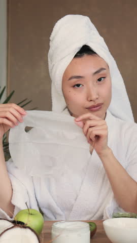 A young Asian woman with a towel wrapped around her head applies a hydrating sheet mask to her face for skincare