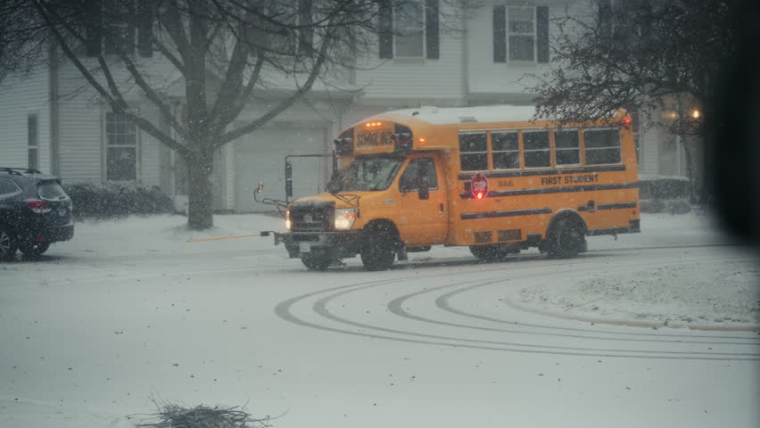 Yellow school bus stopping at suburban neighborhood during active snowfall. Snow blankets the street as flakes fall steadily, creating cold winter scene POV View