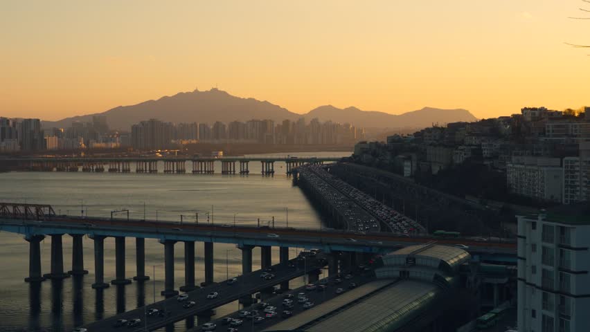 High angle view of busy traffic congestion on Gangbyeonbuk-ro expressway, Dongho and Hannam bridges during golden sunset with Seoul city buildings and mountain silhouettes in South Korea