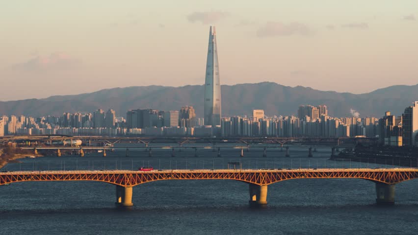 Traffic on parallel bridges over Hangang river with orange Seongsu bridge in foreground and Lotte Tower surrounded by dense apartments and mountains at sunset in Seoul South Korea - aerial close-up