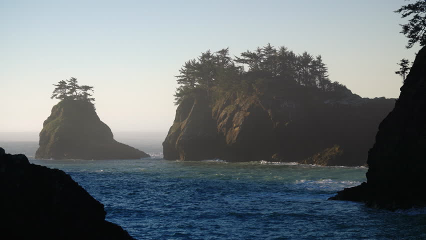 Rocky sea stacks rise from calm ocean waters along Oregon Coast, creating peaceful coastal scene with soft light, depth, and untouched Pacific shoreline atmosphere.