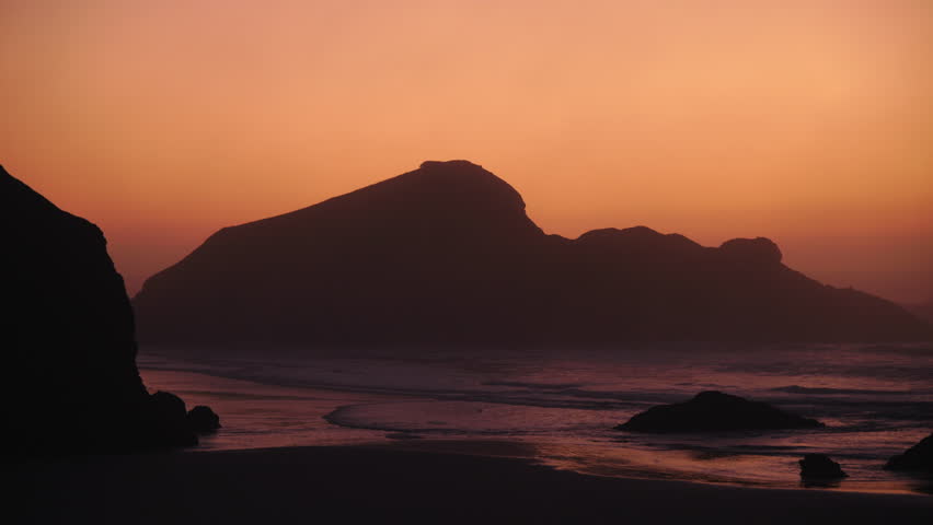 Rocky ocean coastline with waves rolling toward shore under golden sunset sky, creating dramatic seascape, soft reflections, and calm evening coastal atmosphere. Oregon