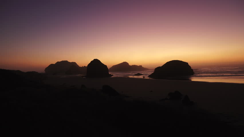 Colorful sunset over rocky ocean beach with waves and dark silhouettes of rocks, creating dramatic coastal scene, calm evening atmosphere, and natural seaside beauty. Oregon USA 