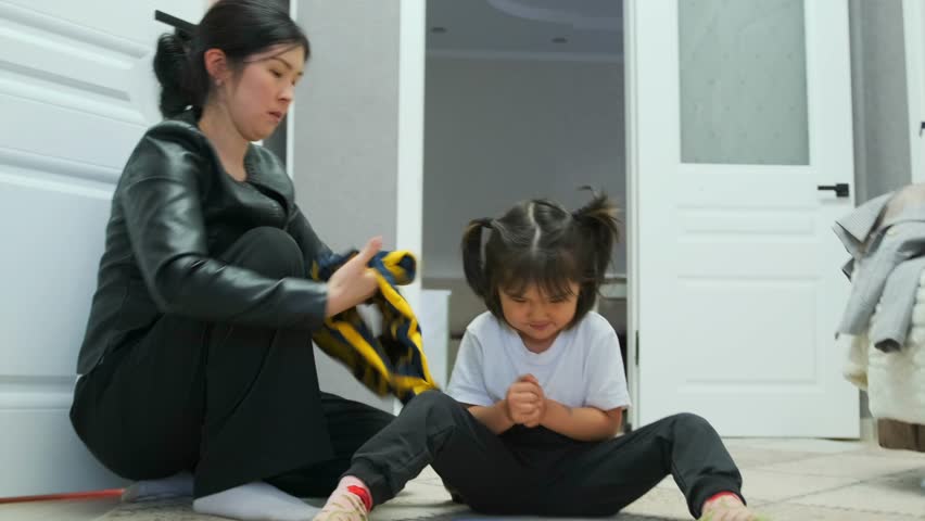Caring Asian mother helps her young daughter put on a striped sweater at home. Morning family routine, motherhood, warmth, and everyday moments of parent-child bonding in domestic atmosphere.