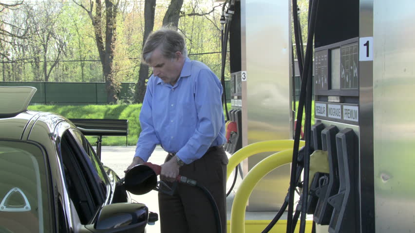 Medium long shot of a man getting out of his car, pumping gas, then checking his