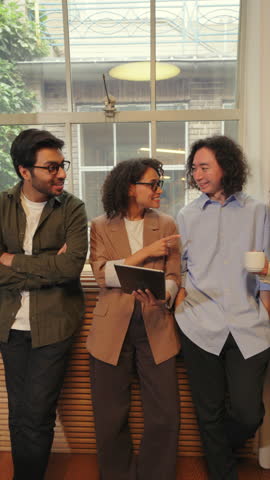 Diverse business people stand by a large window in a modern office, talking openly and sharing ideas while building trust, collaboration, and strong professional connections.