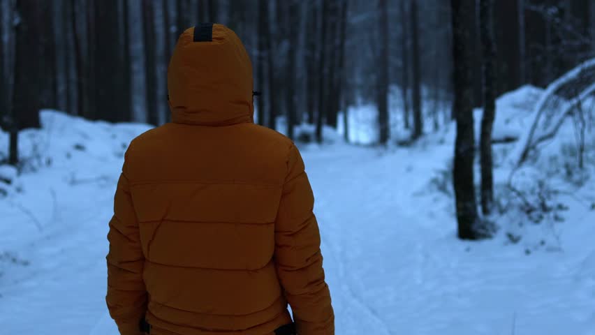 4K video of a man standing in a winter forest in the evening, looking back and then slowly turning toward the camera. The scene is atmospheric and cinematic, with low light, snow-covered trees, and a calm winter mood.