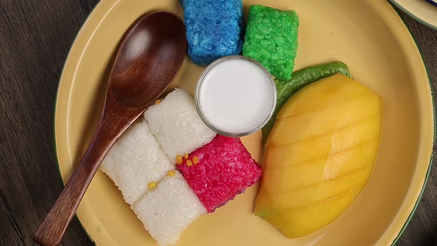 Thai sweet desert red blue yellow white mango sticky rice with coconut cream milk on enamel tin metal plate wood spoon rustic wooden background pan rotate zoom out