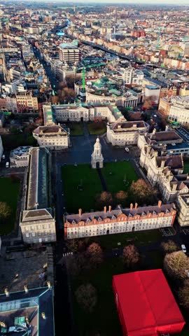 Portrait panning drone shot over Trinity College campus in Dublin at winter sunrise, revealing historic courtyards, stone buildings, frosted lawns, long morning shadows, light traffic nearby