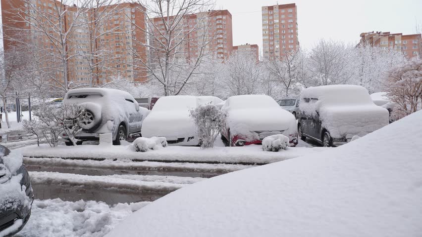 The pavement and vehicles are entirely blanketed in snow in the vicinity of multi-story residential structures. Winter in the urban area.