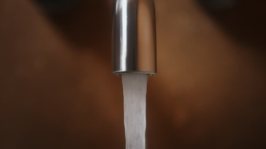 Close-up shot of modern bathroom faucet, bath mixer made of brushed stainless steel. Clear water flows vertically, then human hand turns off the tap. Warm brown wall ceramic tiles background