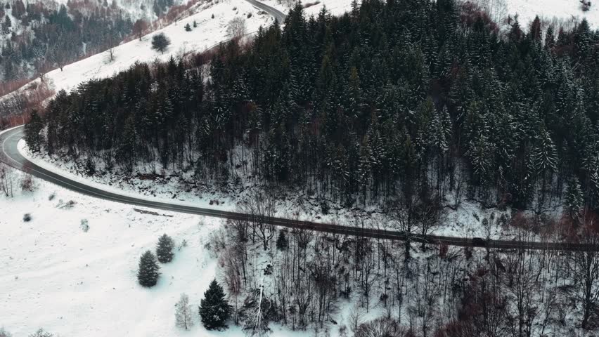 Drone view revealing a curvy road winding through a snow-covered mountain landscape with pine forests