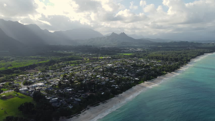 Aerial view of coastal town with white sand beach and turquoise ocean, scenic tropical landscape with green mountains and clouds during vacation