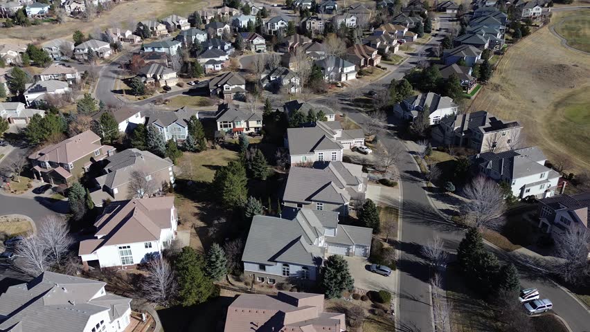 Detached homes near Saddle Rock Golf Club follow winding streets and cul-de-sacs with visible garages, parked vehicles. Roof textures and tree shadows blend into open fairways, Aurora, Colorado. USA