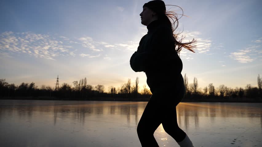Female skater shod in figure skates sliding on ice surface during beautiful sunset. Sportswoman skating on frozen river. Silhouette of woman training at lake. Shining sun reflecting on surface of ice