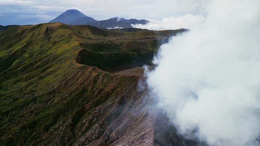 Drone footage capturing Mount Bromo in East Java with crater smoke, rugged volcanic terrain and Mount Semeru visible in the background across the caldera landscape and surrounding mountain ridges.