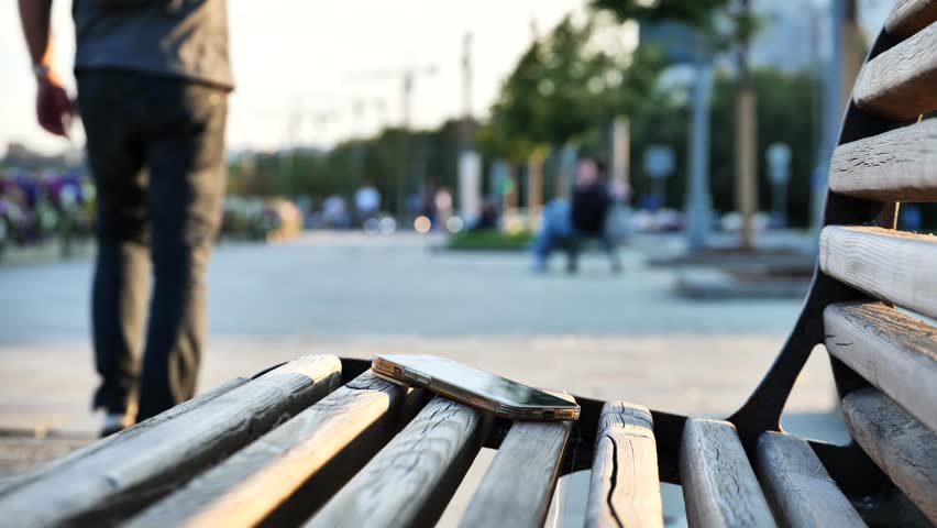 Man bending down to pick up a forgotten phone from wooden park bench