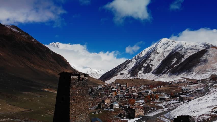 drone view of stone watchtower overlooking village and snowcapped peaks, dramatic clouds casting shadows over steep valley, winding road and river threading through settlement, rugged alpine textures