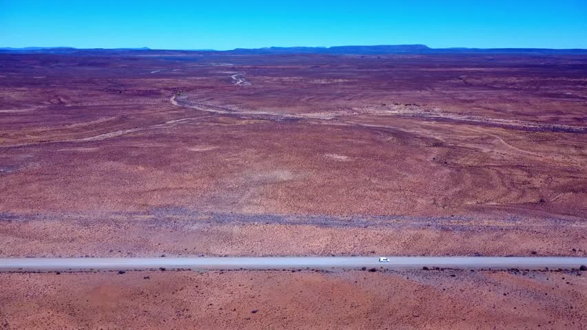 High-angle drone shot capturing a solitary white van traveling along a straight, dusty road through the vast and arid landscape of the Karoo. In the middle of nowhere visualised.