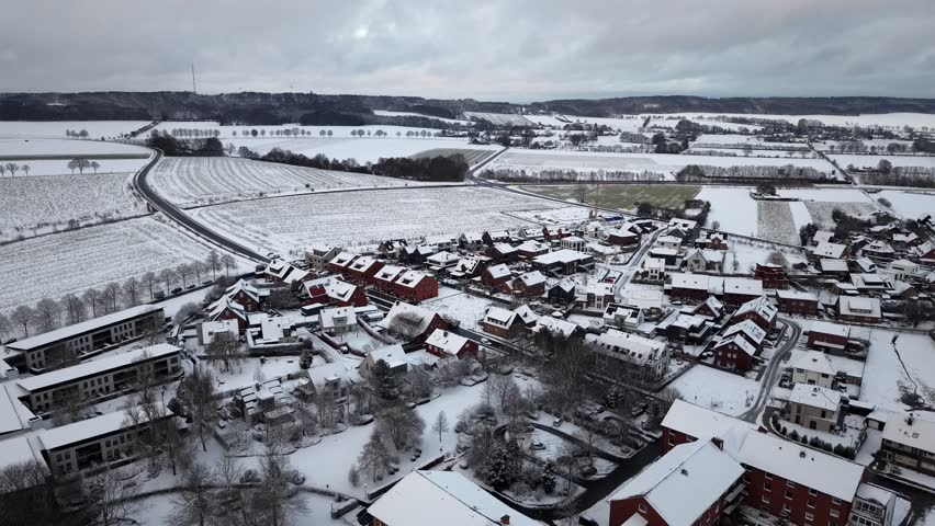 Aerial view of snow-covered rural village surrounded by frozen fields and farmland, with houses, winding roads and winter landscapes on cloudy winter day. Wide shot. Suburb district of town.