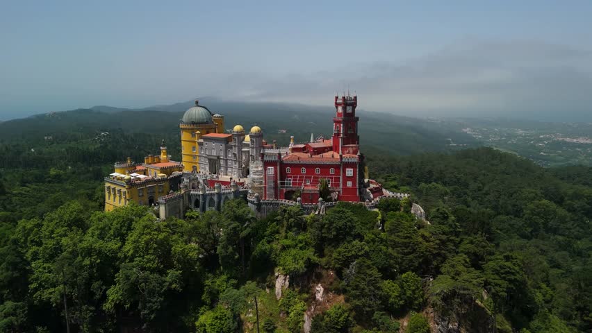Drone approaches then orbits left around Pena Palace in Sintra, Portugal, above forested hills and distant valley views
