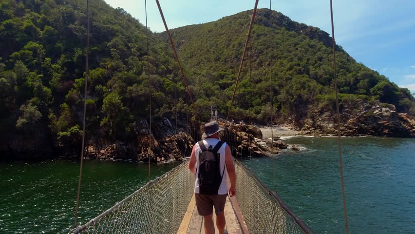 A following shot of a hiker with a backpack walking across the famous suspension bridge in Tsitsikamma National Park, South Africa. 4K.